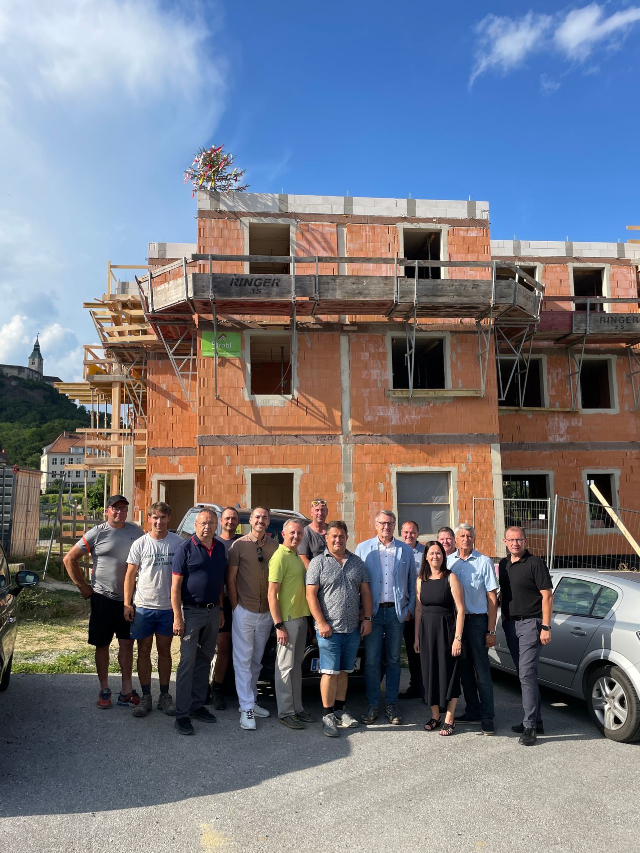 Eine Gruppe von Menschen vor einem im Bau befindlichen Gebäude. Einige tragen Hüte und Brillen, und es sind Autos geparkt. Das Gebäude hat Gerüst und ist aus Ziegeln gebaut.