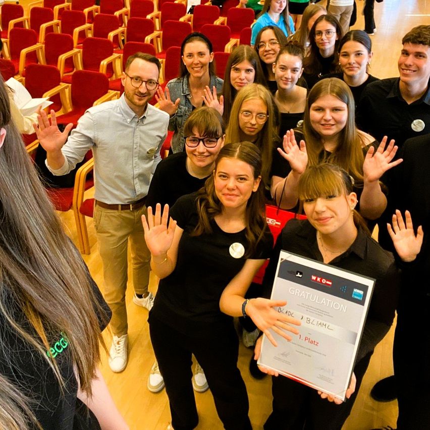 Eine Gruppe von Menschen in schwarzen Shirts hält eine Plakette mit der Aufschrift GRATULATION in einem Saal. Einige tragen Brillen und einige winken. Eine Person in einem blauen Hemd steht hinter ihnen.