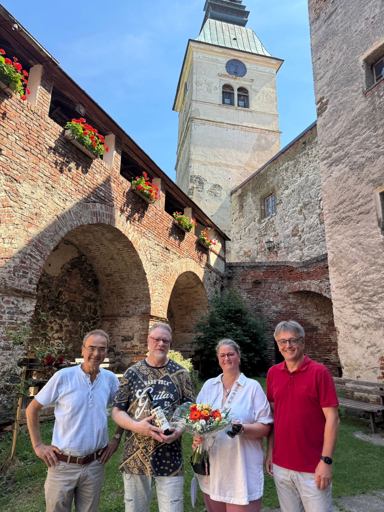 Vier Personen stehen in einem alten Innenhof, einer hält einen Blumenstrauß, ein anderer eine Flasche. Dahinter ist ein Ziegelbogen und ein Turm mit einer Uhr zu sehen.
