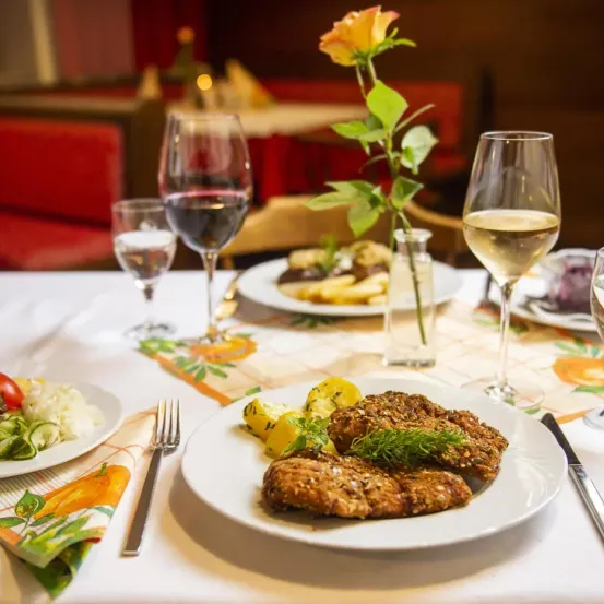 A table set with plates of food, wine glasses, and a flower in a vase. The food includes meat, vegetables, and bread.