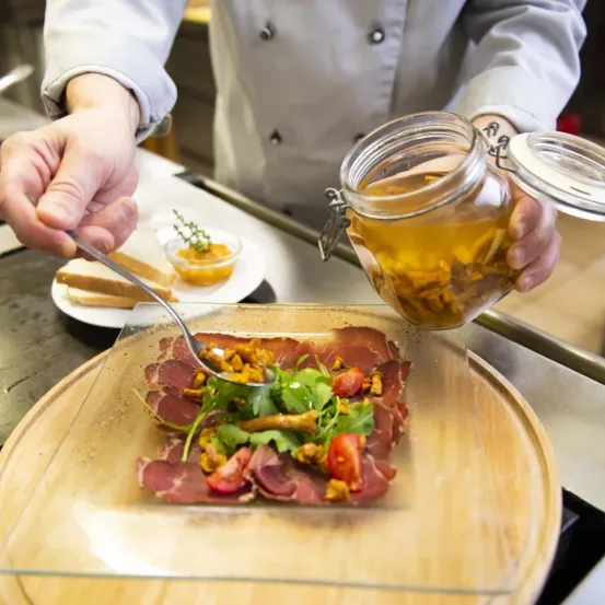 A chef in a kitchen is preparing a dish with sliced meat, vegetables, and a dressing on a wooden plate. Nearby, there is a jar with a liquid inside.