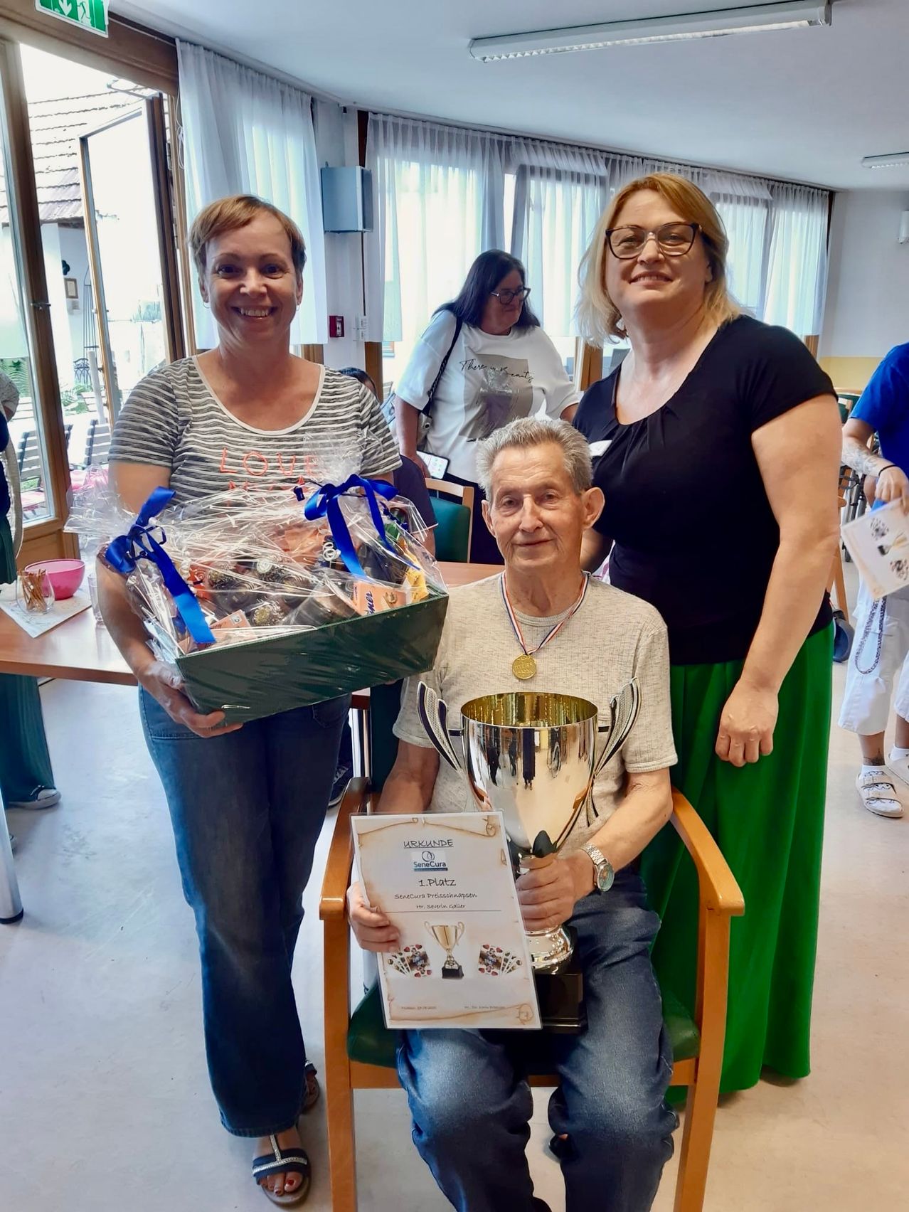 Ein älterer Mann mit einer Medaille sitzt in einem Stuhl und hält einen Pokal. Zwei Frauen stehen hinter ihm, eine hält eine Geschenkbox. Sie sind drinnen, in der Nähe eines Tisches mit Gegenständen darauf. Eine Frau im Hintergrund trägt eine Brille.