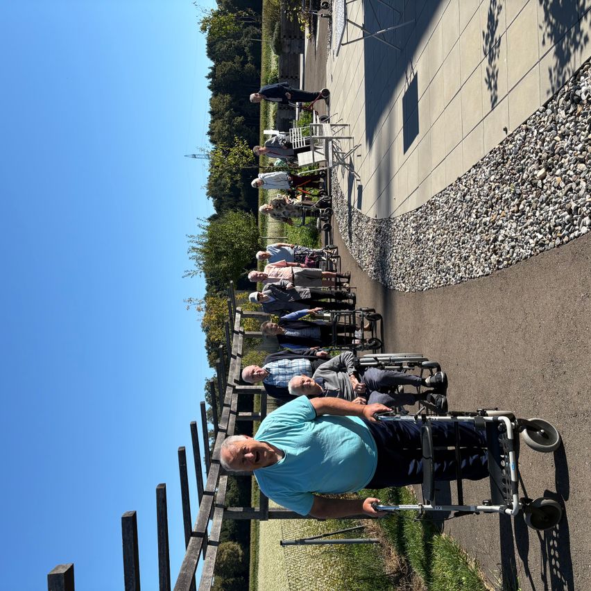 Eine Gruppe älterer Menschen geht mit ihren Rollstühlen in einer Reihe, auf einem Weg, der von Holzgittern gesäumt ist. Im Hintergrund sind Bäume und ein klarer blauer Himmel zu sehen.