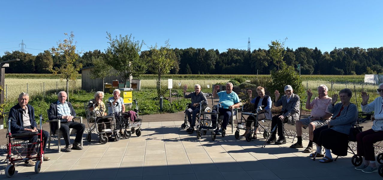Mehrere ältere Menschen sitzen in Rollstühlen in einem Garten im Freien und winken. Dahinter stehen Bäume, ein Feld und ein Turm in der Ferne.