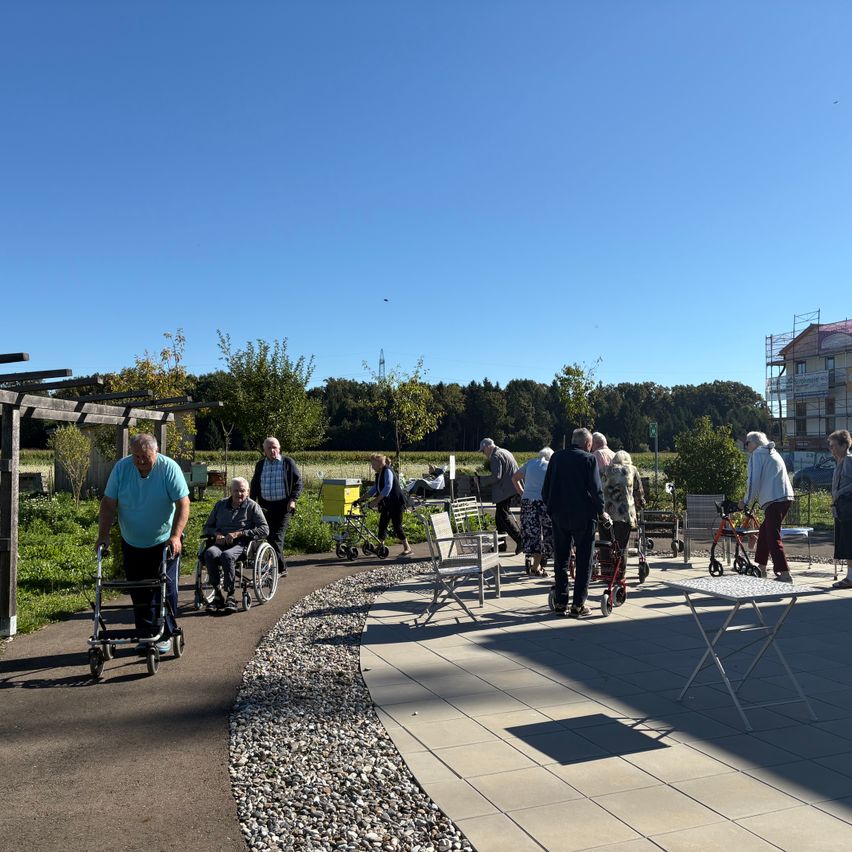 Ein Außenbereich mit älteren Erwachsenen, die auf einem gepflasterten Weg gehen. Bäume und Pflanzen umgeben den Bereich, und ein im Bau befindliches Gebäude ist im Hintergrund zu sehen.