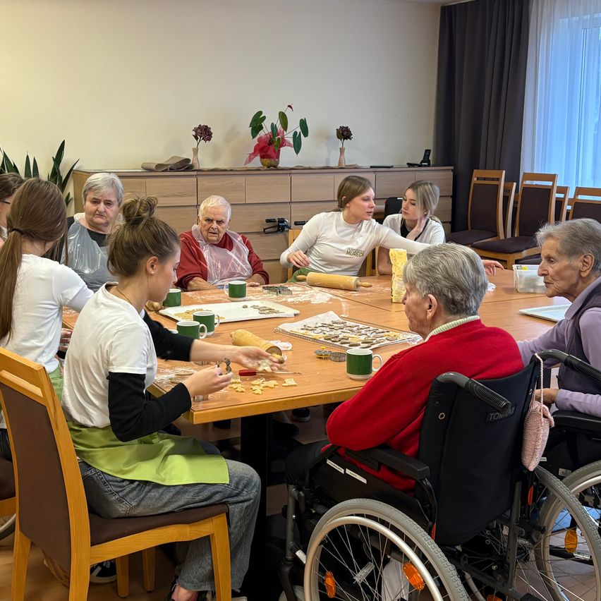 Ältere Frauen und Mädchen sind um einen Tisch versammelt und beschäftigen sich mit einer Backaktivität. Sie haben verschiedene Backwerkzeuge und Zutaten auf dem Tisch. Zwei ältere Frauen sind in Rollstühlen, und eine hält eine Teigrolle.