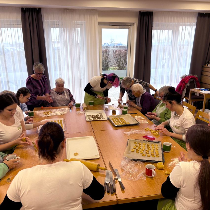 Eine Gruppe älterer Frauen in einer Küche bereitet Essen zu. Sie sitzen um einen Tisch herum mit verschiedenen Lebensmitteln und Utensilien.