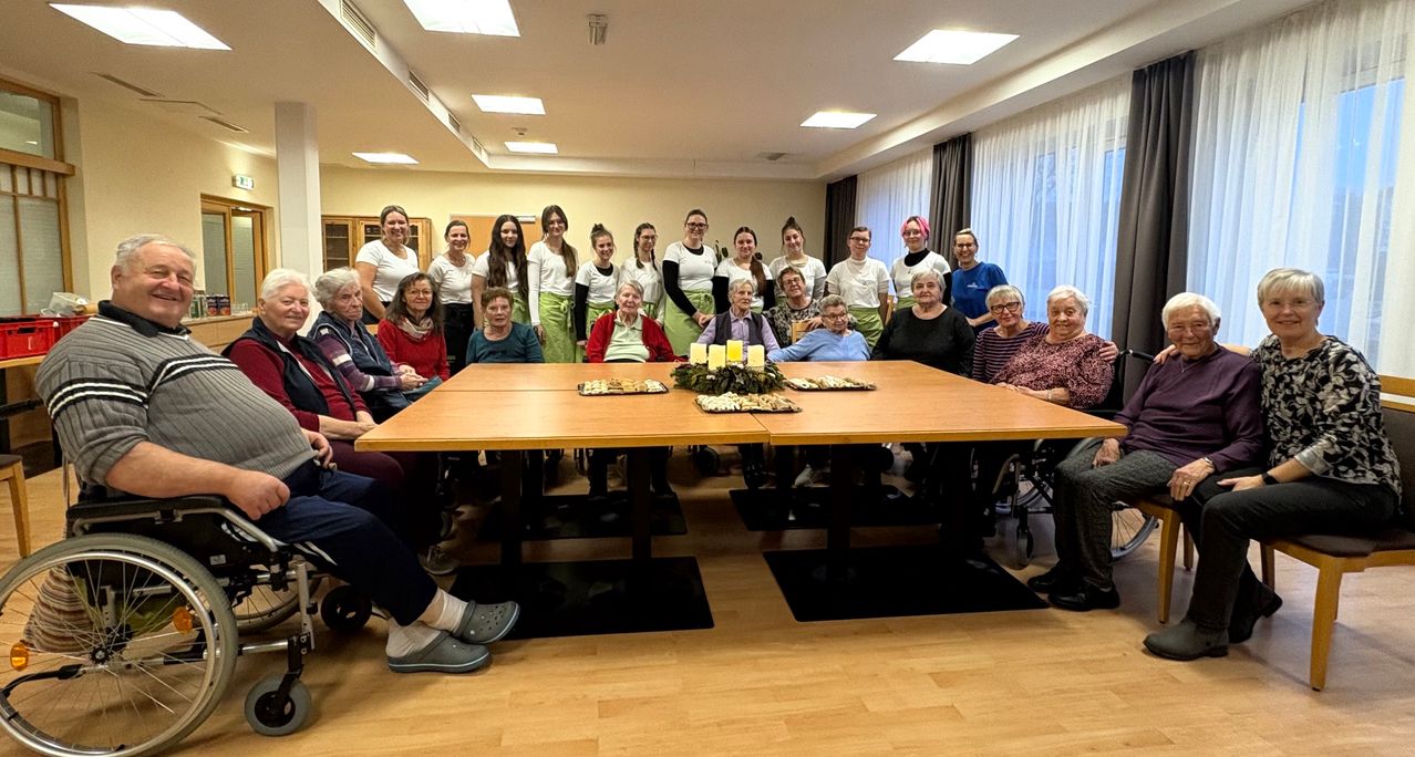 Ein Gruppenfoto von älteren Frauen und jungen Frauen, möglicherweise Studenten, die sich um einen langen Tisch mit Kerzen und Tellern mit Essen versammelt haben. Der Raum hat Holzböden und weiße Vorhänge.