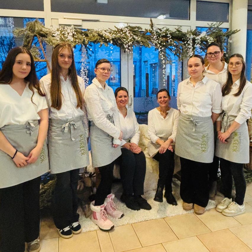 Eine Gruppe von Frauen in weißen Hemden und grauen Schürzen posiert für ein Foto in einem mit Weihnachtsdekoration geschmückten Raum. Zwei Frauen sitzen im Hintergrund auf einem Stuhl.