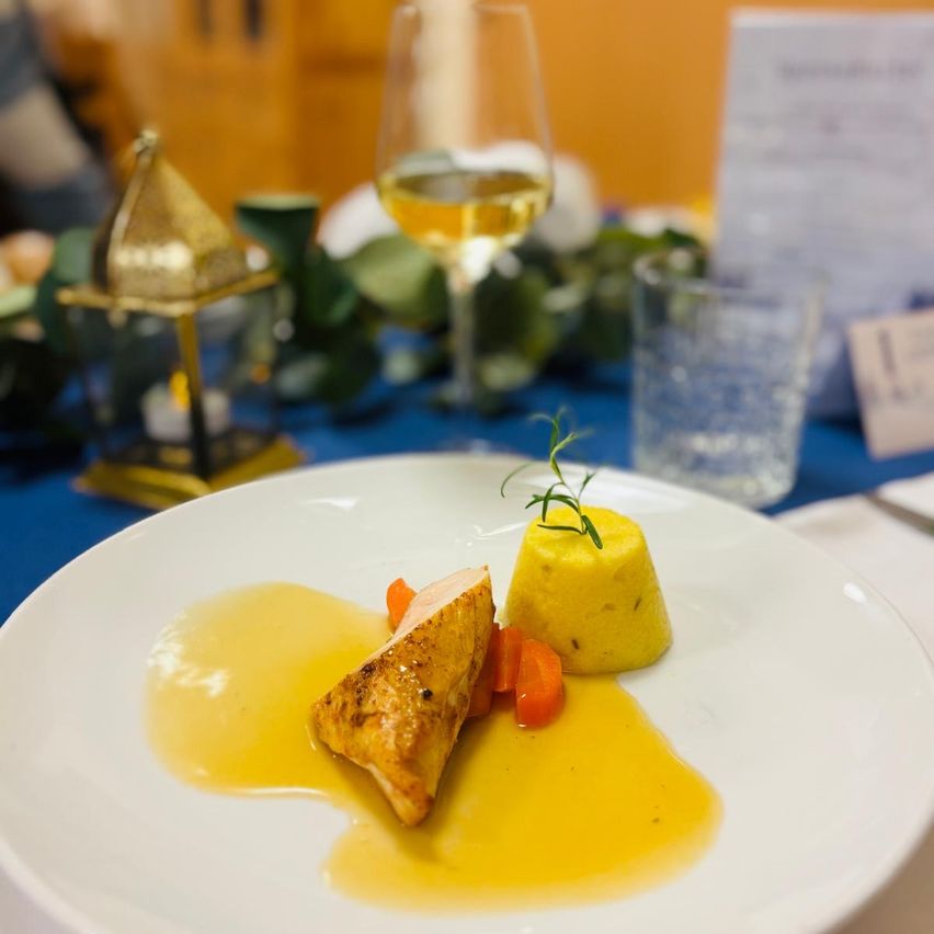 Ein weißer Teller mit einem Gericht aus Hähnchen, gelber Soße und einer Beilage von Karotten, garniert mit einem Rosmarinzweig. Im Hintergrund ein Glas Weißwein und ein blauer Tischläufer.