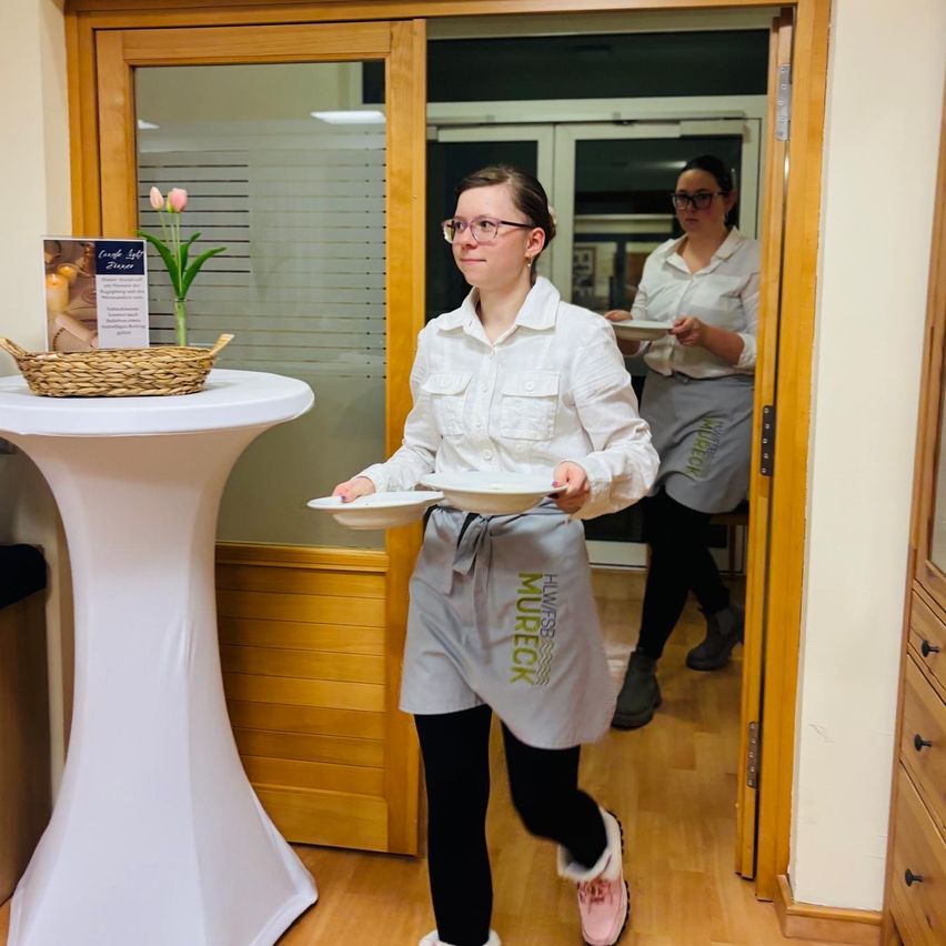 Zwei Frauen, beide in Schürzen, gehen mit Tellern durch eine Holztür. Eine von ihnen schaut nach links, während die andere nach vorne schaut. Ein Tisch mit einem Korb und Blumen steht auf der linken Seite.