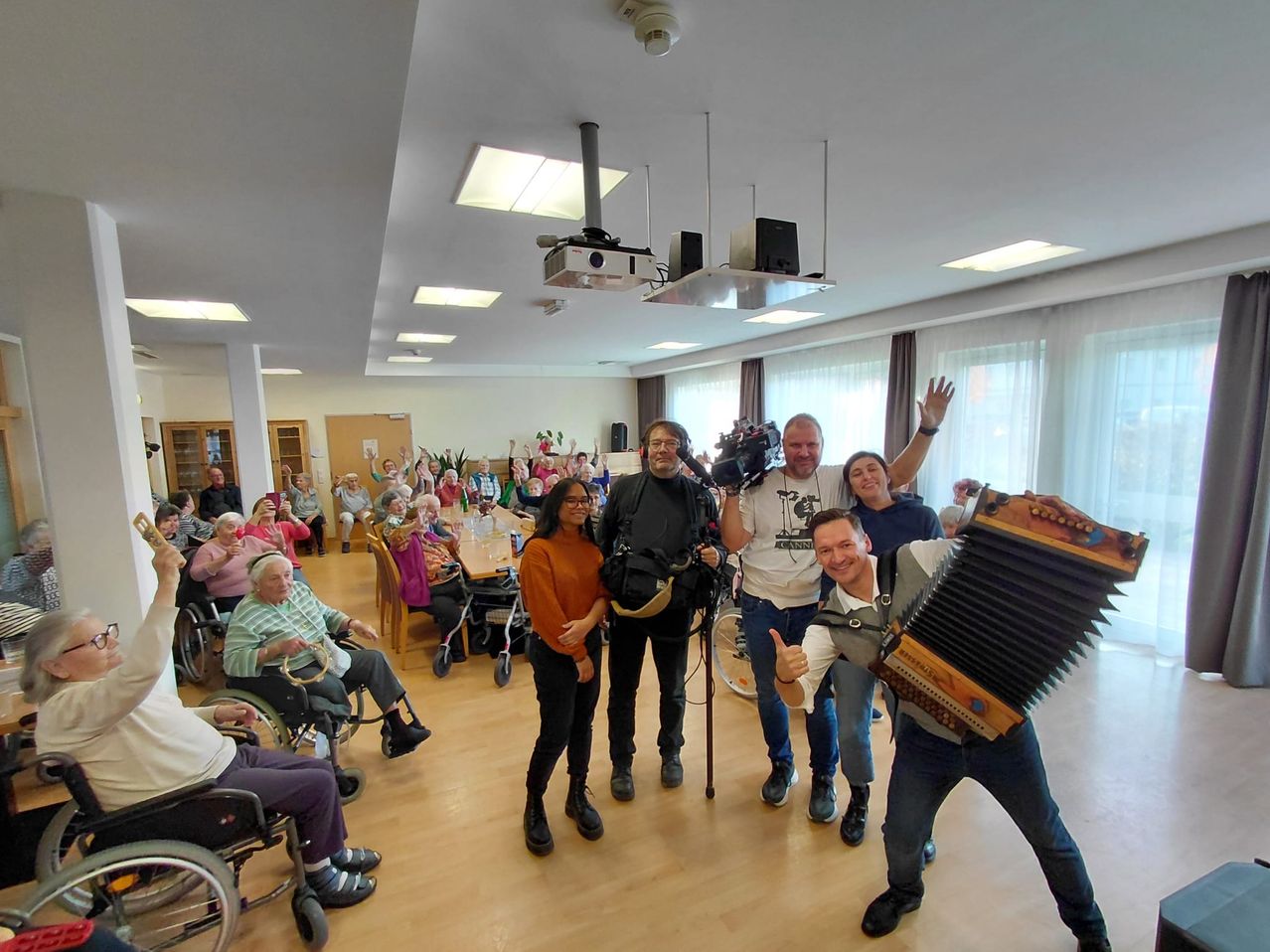 Eine Gruppe von Menschen in einem Raum, einige sitzen in Rollstühlen, einer hält eine Kamera und ein anderer spielt Akkordeon. Mehrere andere lächeln und heben ihre Hände.