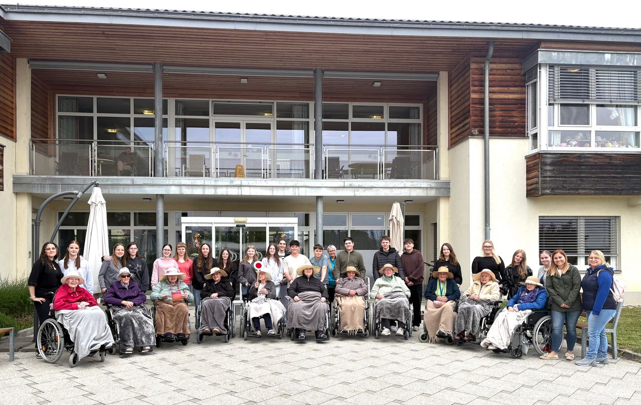 Eine Gruppe älterer Menschen in Rollstühlen und junge Personen posieren für ein Foto vor einem Gebäude mit Glaswänden und einer Terrasse.