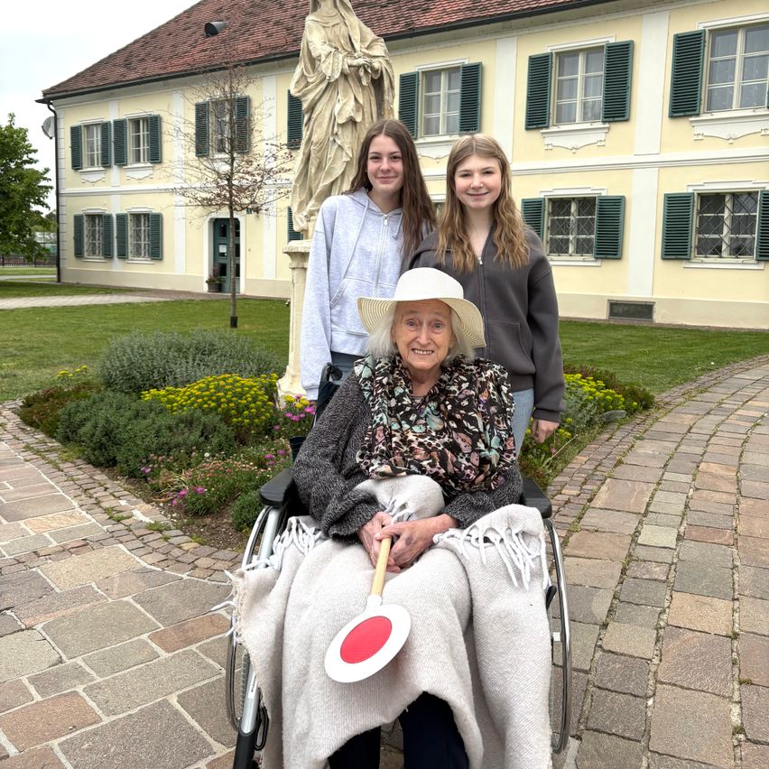 Eine ältere Frau in einem Rollstuhl mit einer Decke hält eine Paddel. Zwei Mädchen stehen hinter ihr. Sie stehen vor einem Gebäude mit einer Statue.
