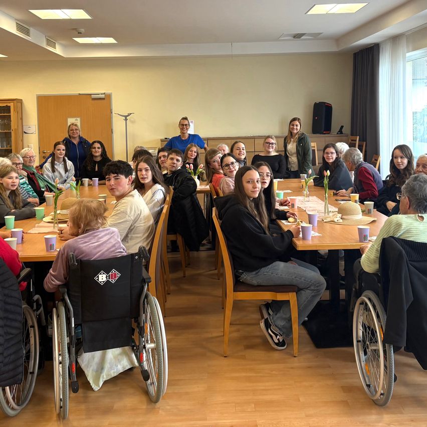 Eine Gruppe von Menschen, einschließlich einiger in Rollstühlen, sitzt um einen Tisch in einem Raum und isst gemeinsam.