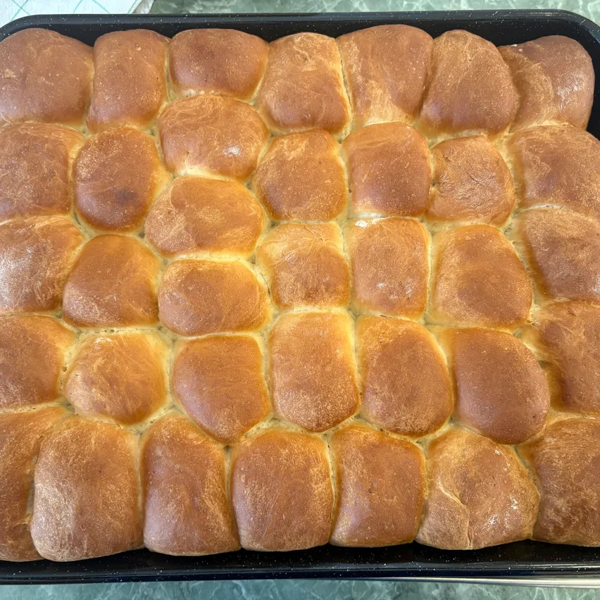 Ein Tablett gefüllt mit goldbraunen, frisch gebackenen Dinner-Rolls, die in einem ordentlichen Rastermuster angeordnet sind. Jeder Brötchen hat eine leicht knusprige Kruste und ein weiches, flauschiges Inneres.