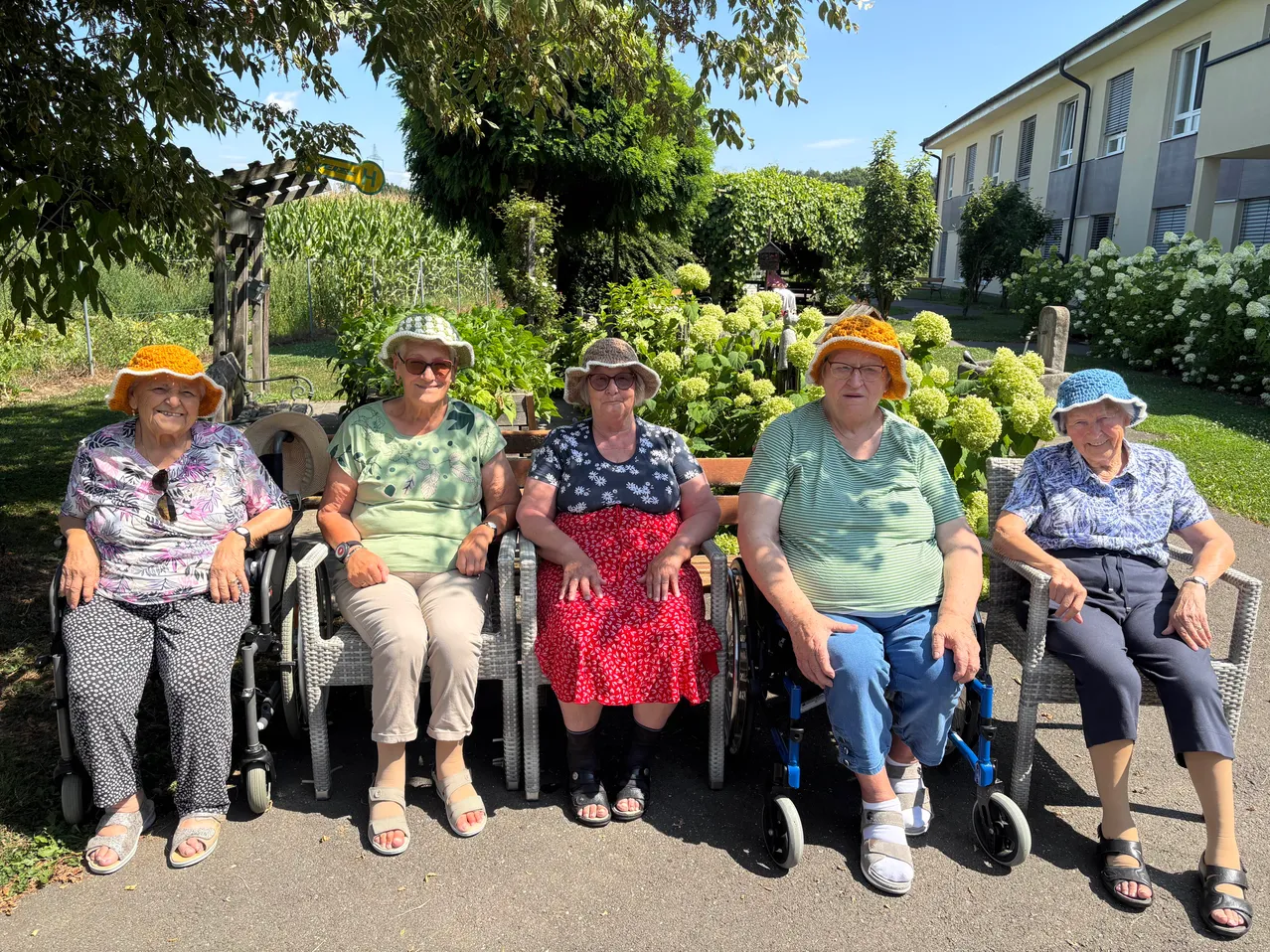 Fünf ältere Frauen in Hüten und Brillen sitzen in Rollstühlen in einem Garten mit üppiger Begrünung und einem Gebäude im Hintergrund.