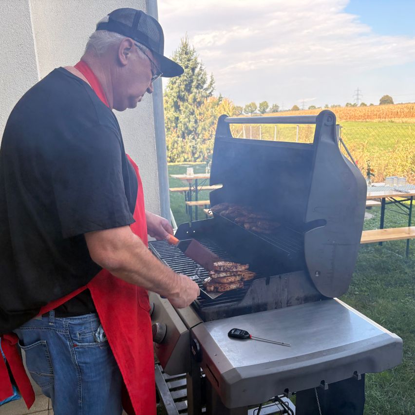An older man wearing a red apron and a hat is grilling meat on a barbecue grill outdoors.