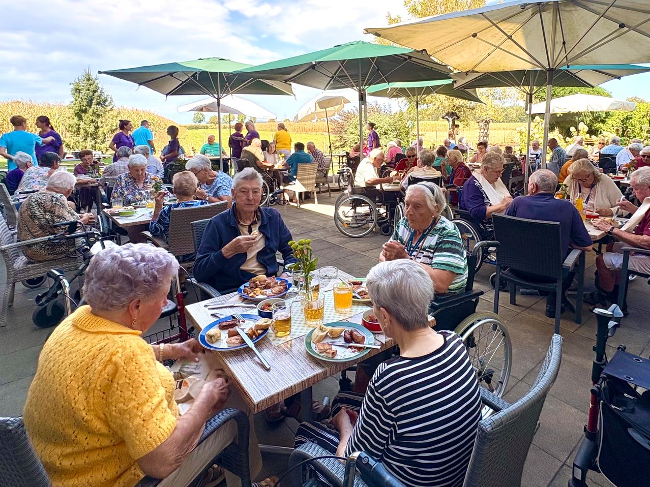 A group of elderly people are dining outdoors at a restaurant with umbrellas and wheelchairs. They are enjoying their meals and chatting.