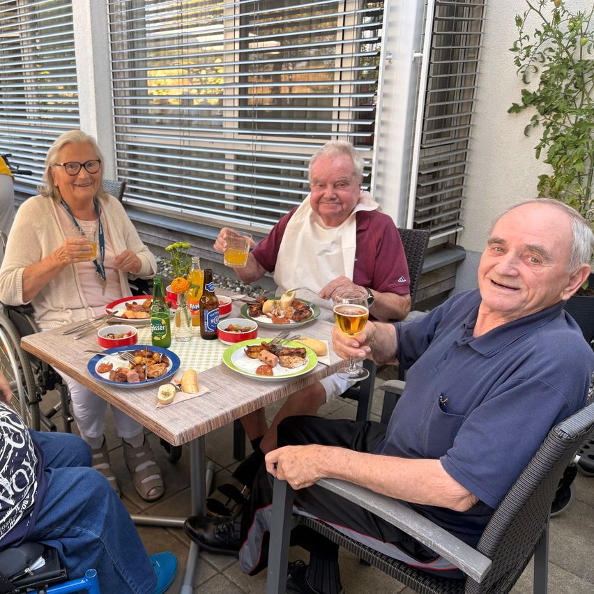 Four elderly people are sitting around a table, smiling and holding drinks. One man is wearing an apron. There are plates of food, bottles, and a plant on the table.