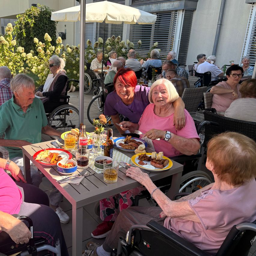 A group of elderly people are sitting around a table in a garden, enjoying their meal. One woman is smiling and holding a fork.