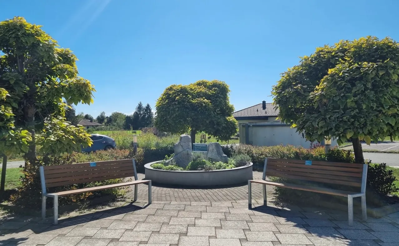 Ein Park mit zwei Bänken, einem runden Garten mit einem Felsen und Pflanzen und einem Haus im Hintergrund.