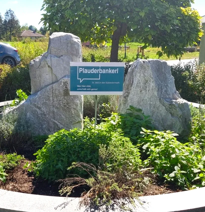 Ein Gartenbereich mit großen Felsen und einem Schild, das Plauderbankerl steht. Der Garten ist von Pflanzen und Bäumen umgeben.