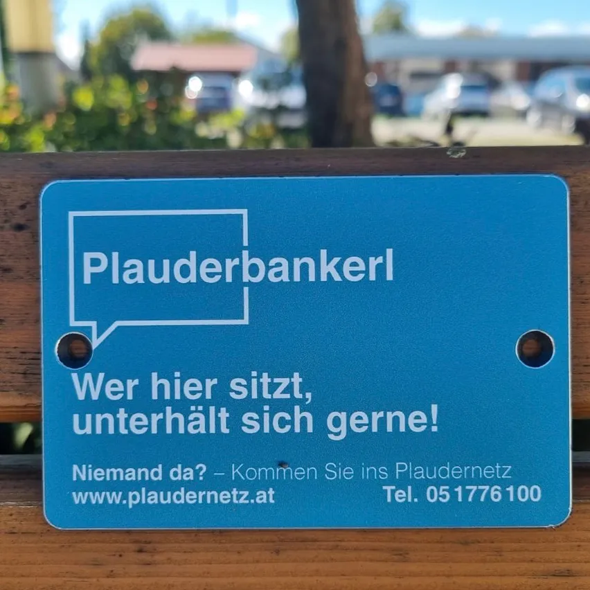 Ein blaues Schild auf einer Holzbank mit dem Text 'Plauderbankerl' und einer Sprechblase. Es lädt dazu ein, dem Plaudernetz beizutreten.