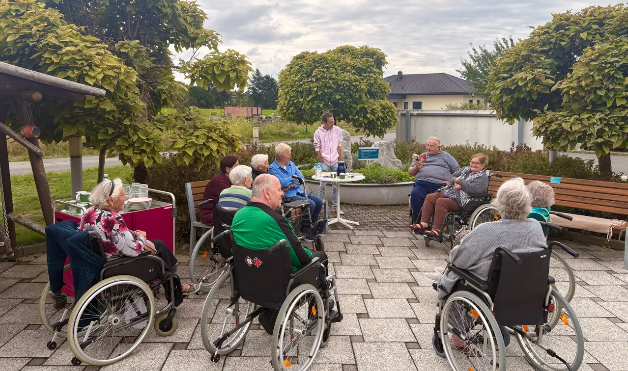 Eine Gruppe älterer Menschen in Rollstühlen versammelt sich in einem Außenbereich, wobei ein Mann steht und ein Buch liest.