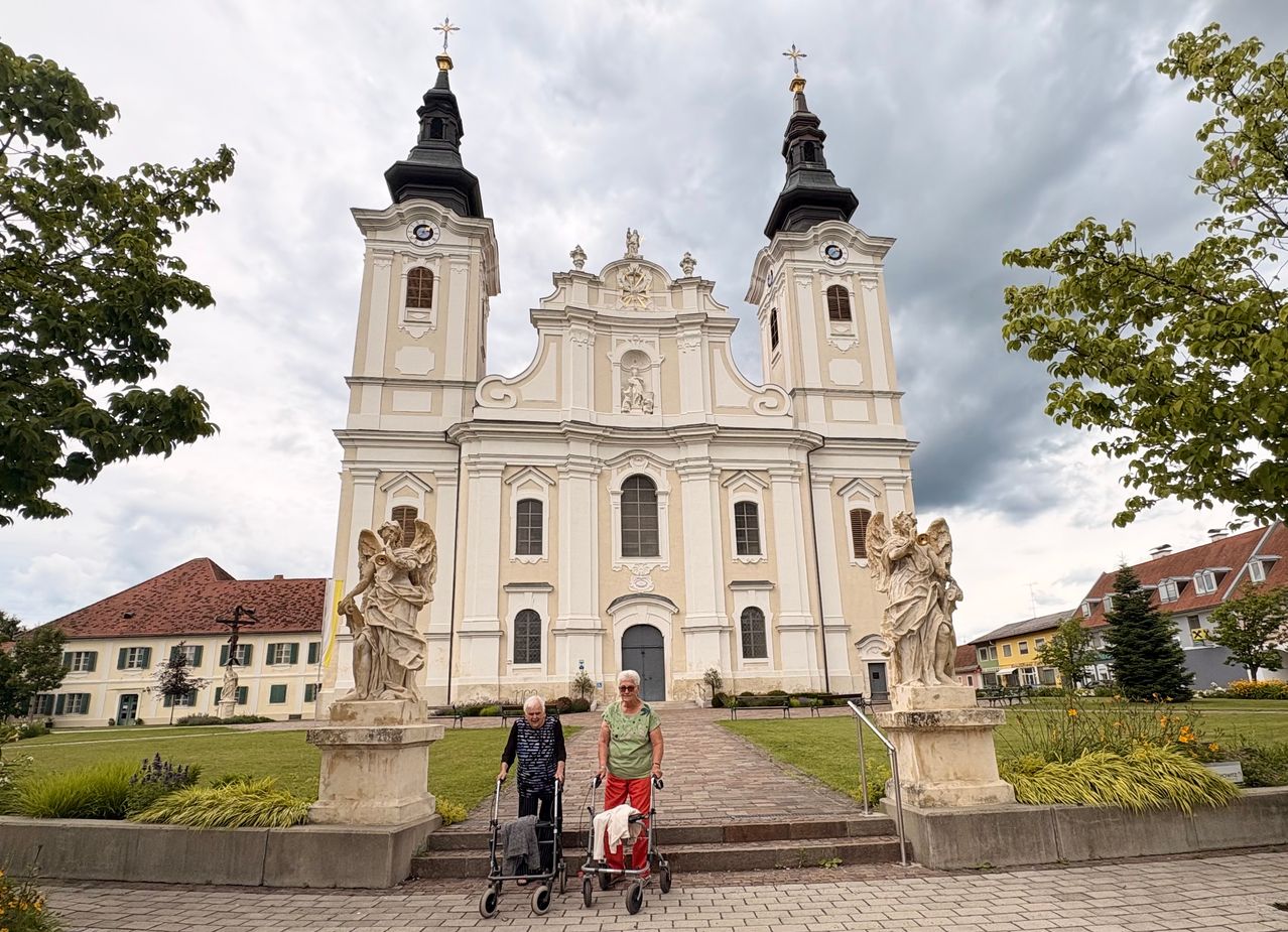 Zwei ältere Frauen gehen mit ihren Rollstühlen vor einer weißen Kirche mit zwei Türmen und Statuen.