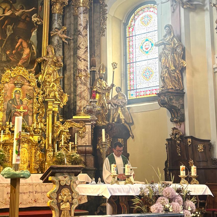 Ein Priester in einer Kirche, der vor einem Altar mit Kerzen steht. Eine goldene Statue steht über ihm.