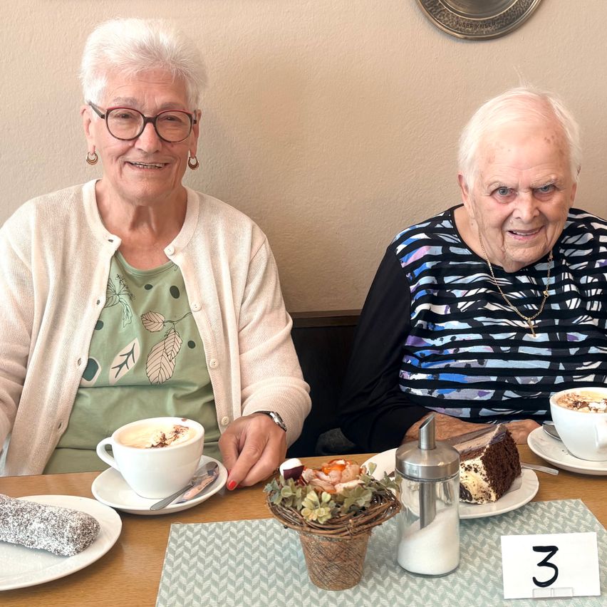 Zwei ältere Frauen sitzen an einem Tisch mit Essen und Getränken, lächeln und schauen in die Kamera. Eine Frau hat eine weiße Strickjacke und die andere eine Halskette.