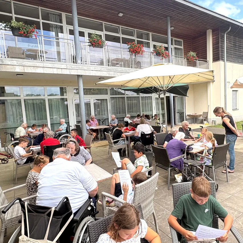 Essensbereich im Freien mit vielen Tischen und Stühlen, mehrere Erwachsene und Kinder sitzen. Sonnenschirme und Blumentöpfe auf dem Balkon. Eine Frau steht in der Nähe der Tische, möglicherweise serviert.
