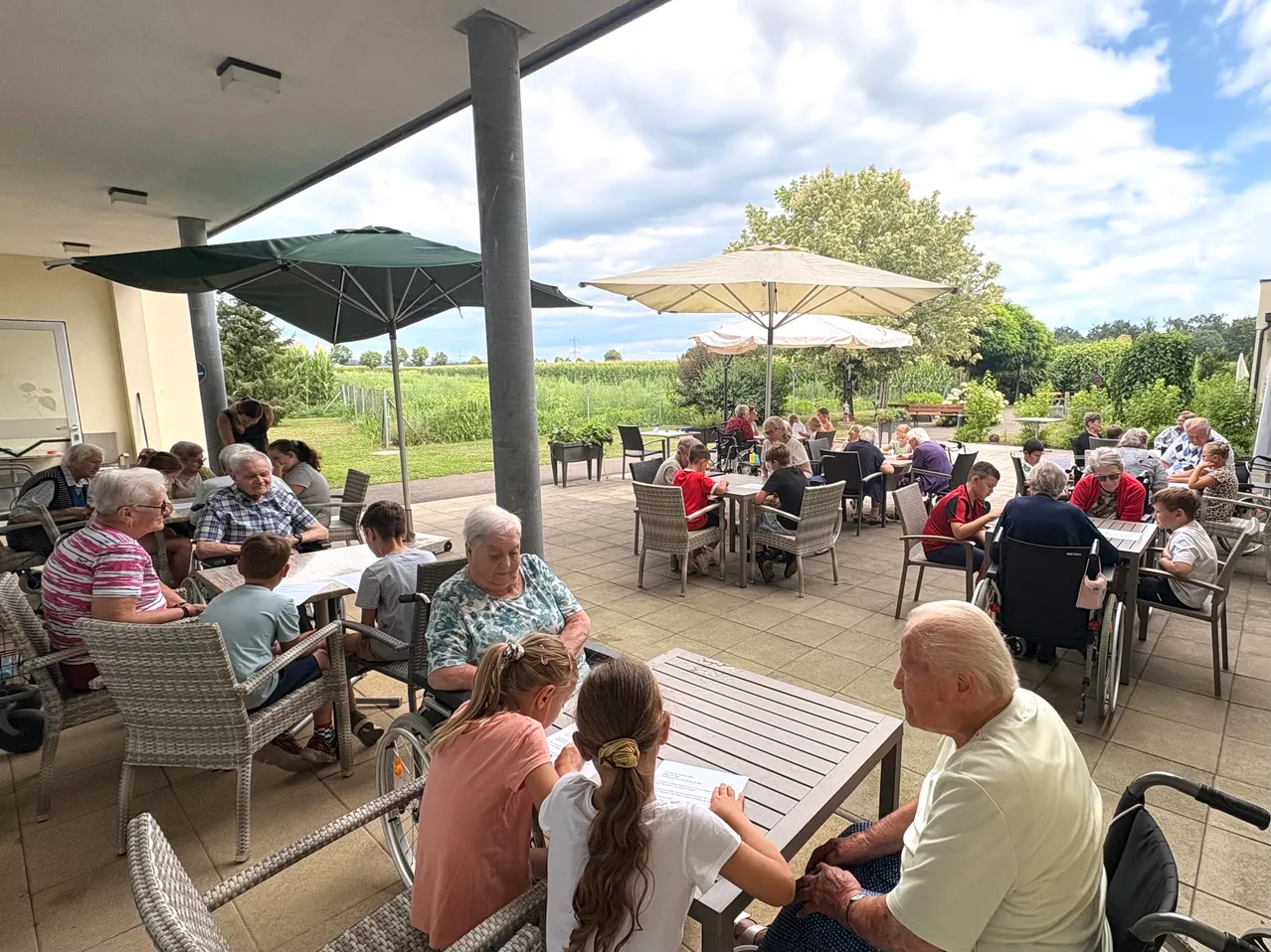 Eine Gruppe älterer Menschen sitzt draußen unter Sonnenschirmen, einige schreiben. Kinder sitzen an einem Tisch. Bäume und ein Feld sind im Hintergrund.