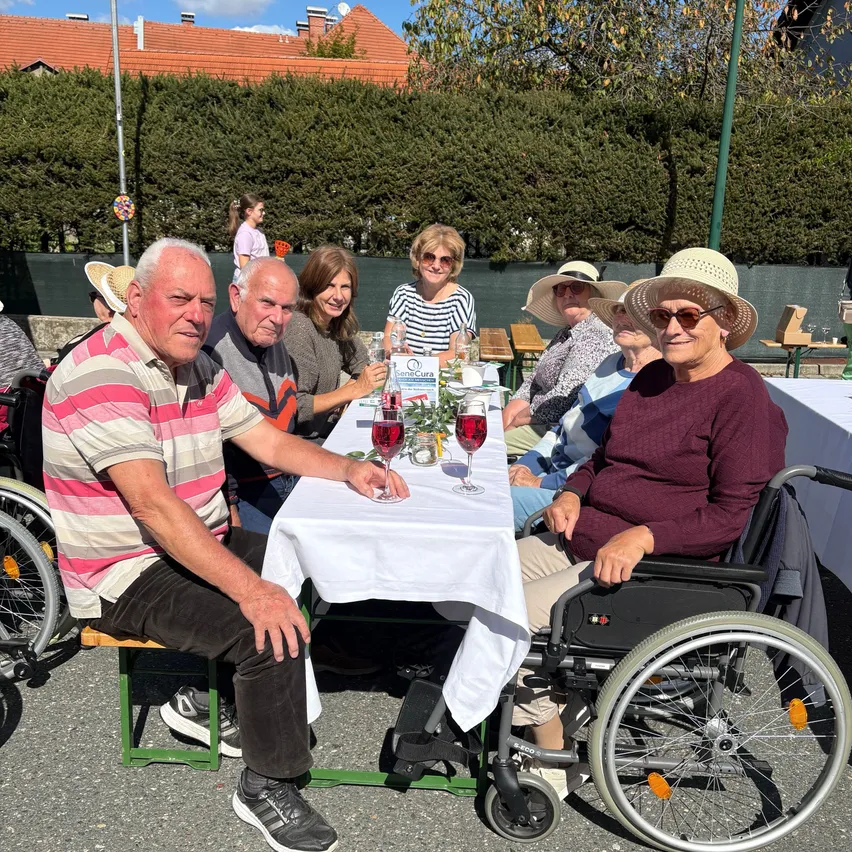 Eine Gruppe älterer Menschen, einige in Rollstühlen, sitzen um einen Tisch herum und genießen eine Mahlzeit im Freien. Der Tisch ist mit einem weißen Tuch bedeckt und es gibt Weinläser und eine Blumenarrangement. Im Hintergrund befinden sich Hecken und ein Gebäude.