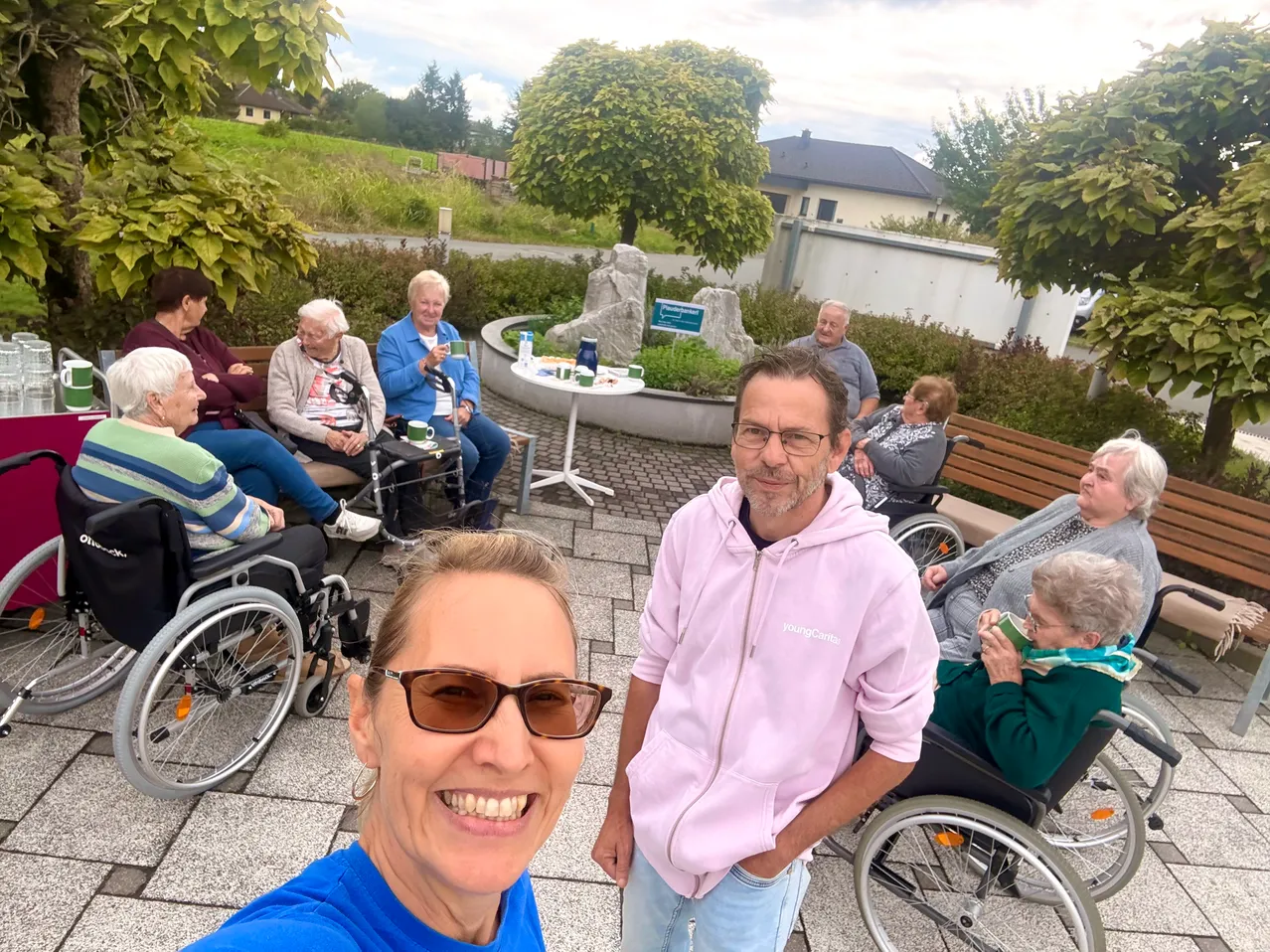Eine Gruppe älterer Menschen sitzt auf Bänken und Rollstühlen in einem Park. Ein Mann und eine Frau stehen vor ihnen und lächeln.