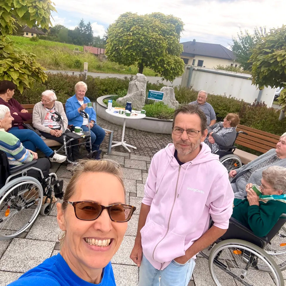 Eine Gruppe älterer Menschen sitzt auf Bänken und Rollstühlen in einem Park. Ein Mann und eine Frau stehen vor ihnen und lächeln.