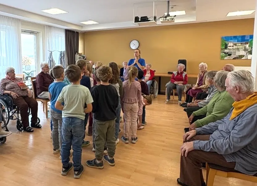 Eine Gruppe von Kindern und älteren Menschen versammelt sich in einem Raum. Eine Frau in einem blauen Shirt steht vor ihnen und hält möglicherweise eine Rede.