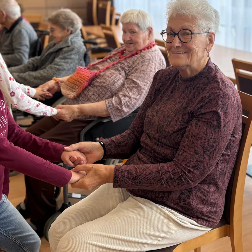Eine ältere Frau mit weißen Haaren sitzt in einem Stuhl und lächelt, während sie die Hand eines jungen Menschen hält. Hinter ihr sitzt eine weitere ältere Frau und lächelt. Sie befinden sich in einem Raum mit Holzböden und Stühlen.