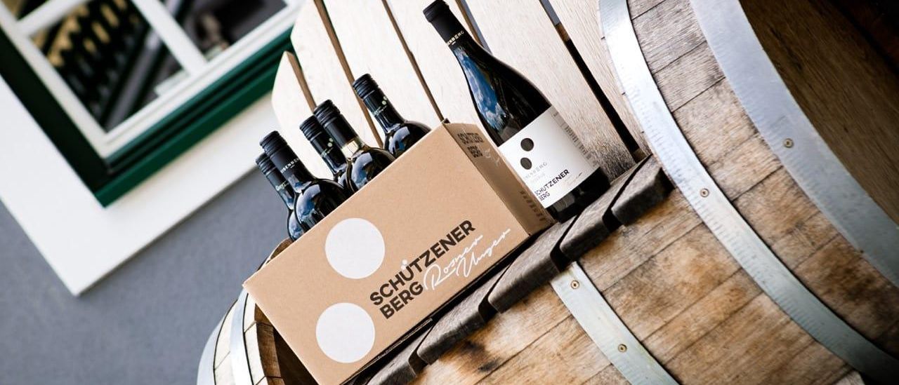 Ein hölzerner Fass mit einer Schachtel, die Schutzener Berg darauf, oben, enthält mehrere Flaschen Wein mit einem sichtbaren Etikett.