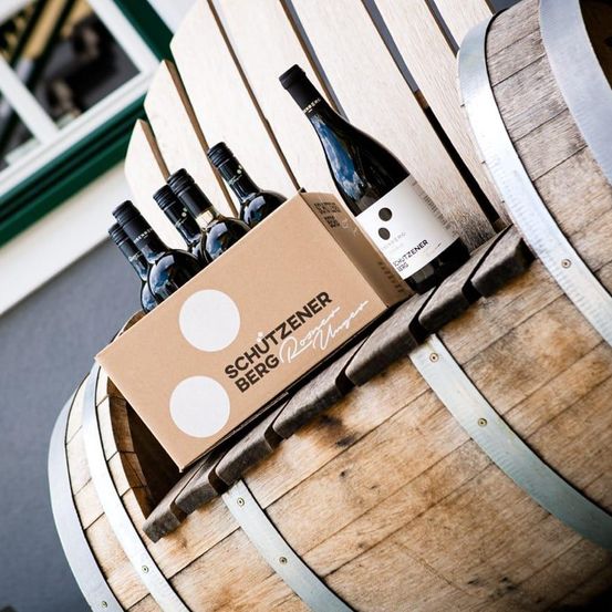 Ein hölzerner Fass enthält eine Schachtel Schutzener Berg Weine. Die Schachtel zeigt weiße Kreise und den Markennamen. Die Weinflaschen sind dunkel mit weißen Etiketten.