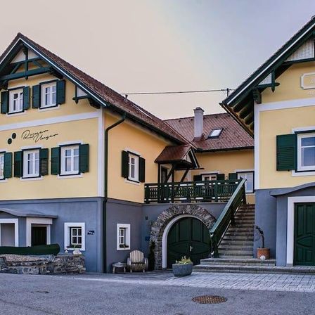 Ein zweistöckiges gelbes Gebäude mit grünen Fensterläden und einem braunen Dach. Es gibt eine Treppe, die zu einem Balkon führt. Draußen gibt es Pflanzen und einen Stuhl. Das Gebäude hat zwei grüne Türen.