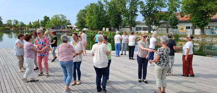 Eine Gruppe von Erwachsenen steht auf einer Holzplattform, einige tanzen, während andere reden. Hinter ihnen befindet sich ein Gewässer, Bäume und ein Gebäude.