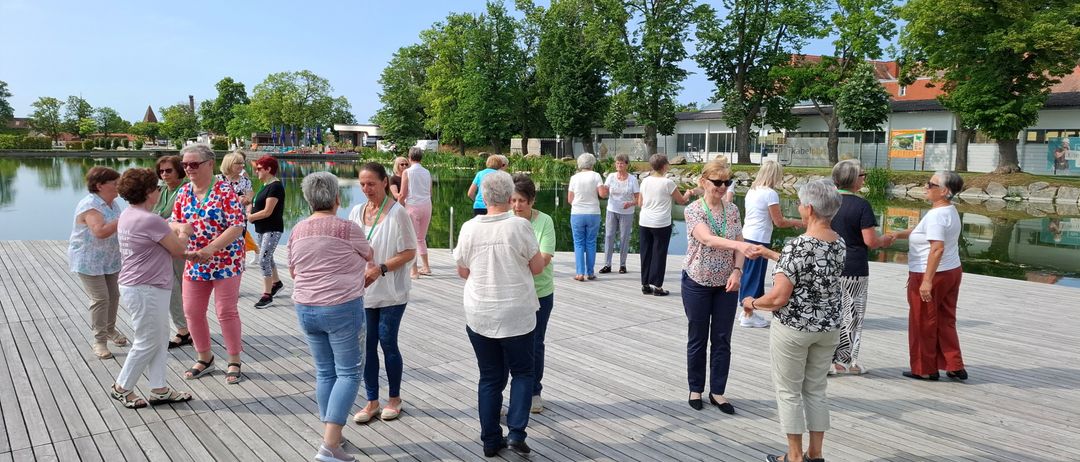 Eine Gruppe von Erwachsenen steht auf einer Holzplattform, einige tanzen, während andere reden. Hinter ihnen befindet sich ein Gewässer, Bäume und ein Gebäude.