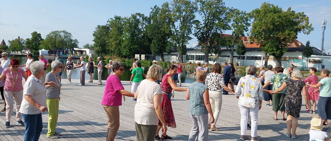 Eine Gruppe von Menschen tanzt auf einer Holzplattform in der Nähe eines Gewässers. Sie halten sich an den Händen in einem Kreis. Das Gebiet ist von Bäumen und Gebäuden umgeben.