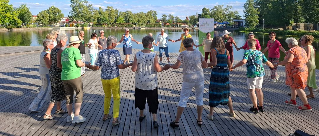 Eine Gruppe von Menschen steht im Kreis, hält sich an den Händen und tanzt auf einer Holzplattform am See.