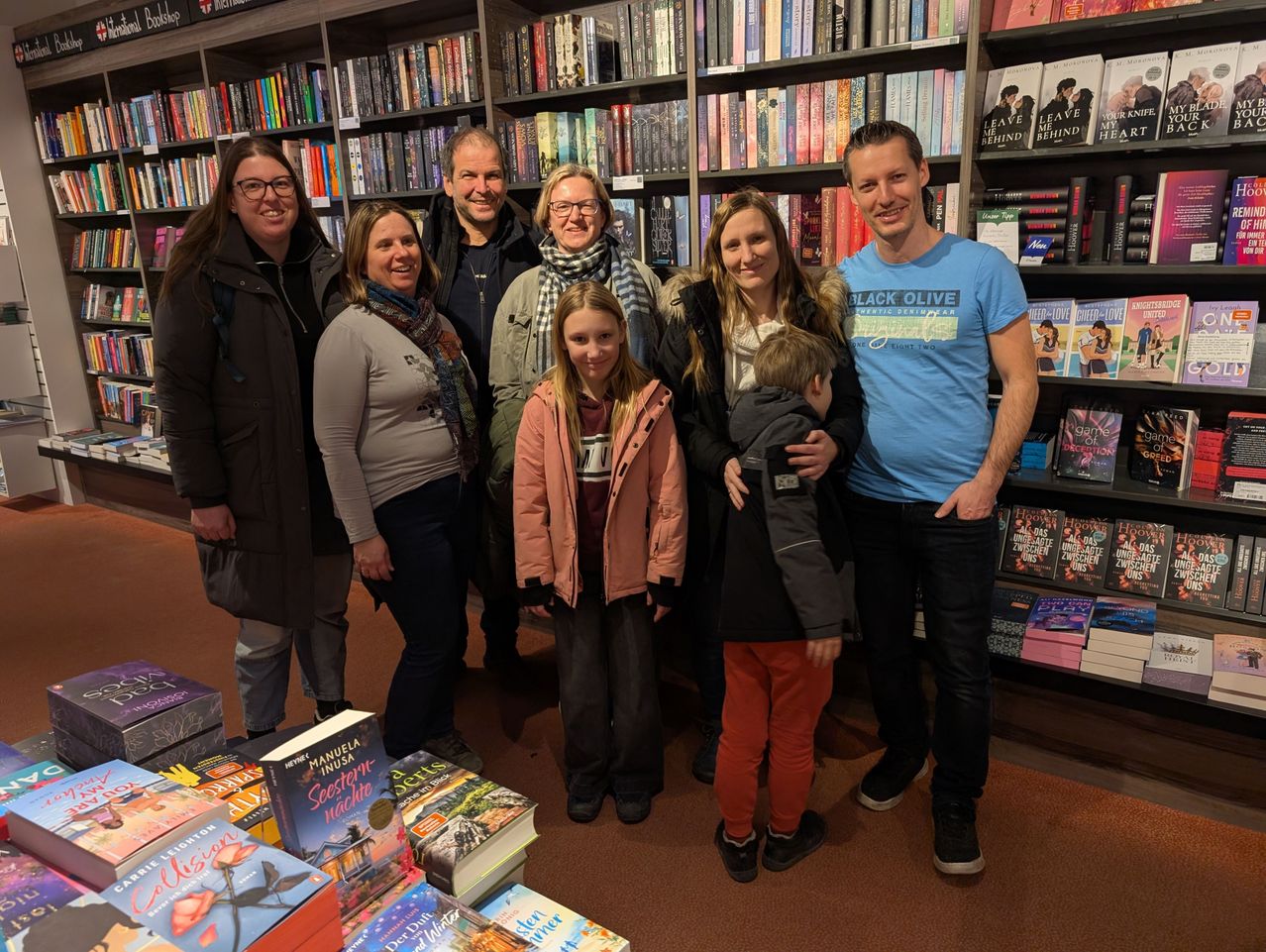 Eine Gruppe von Menschen, einschließlich Erwachsener und Kinder, steht in einem Buchladen zusammen und lächelt für ein Foto. Hinter ihnen sind mit Büchern gefüllte Regale zu sehen.