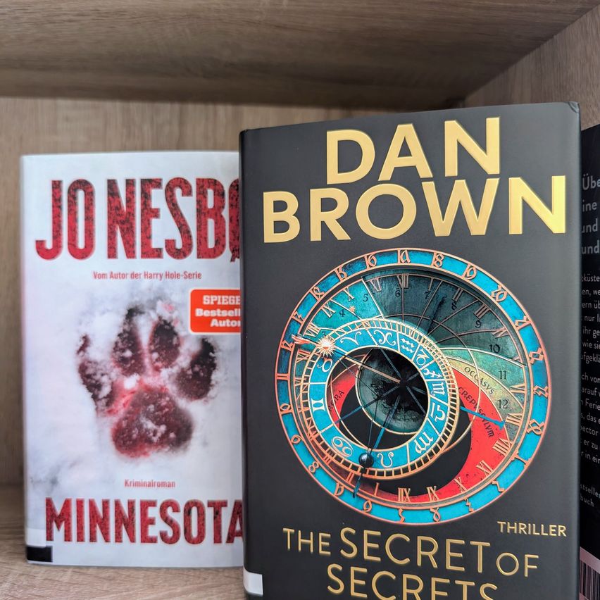 Zwei Bücher stehen auf einem Regal. Eines ist von Dan Brown mit dem Titel 'Das Geheimnis der Geheimnisse' und das andere ist von Jo Nesbo mit dem Titel 'Minnesota'.