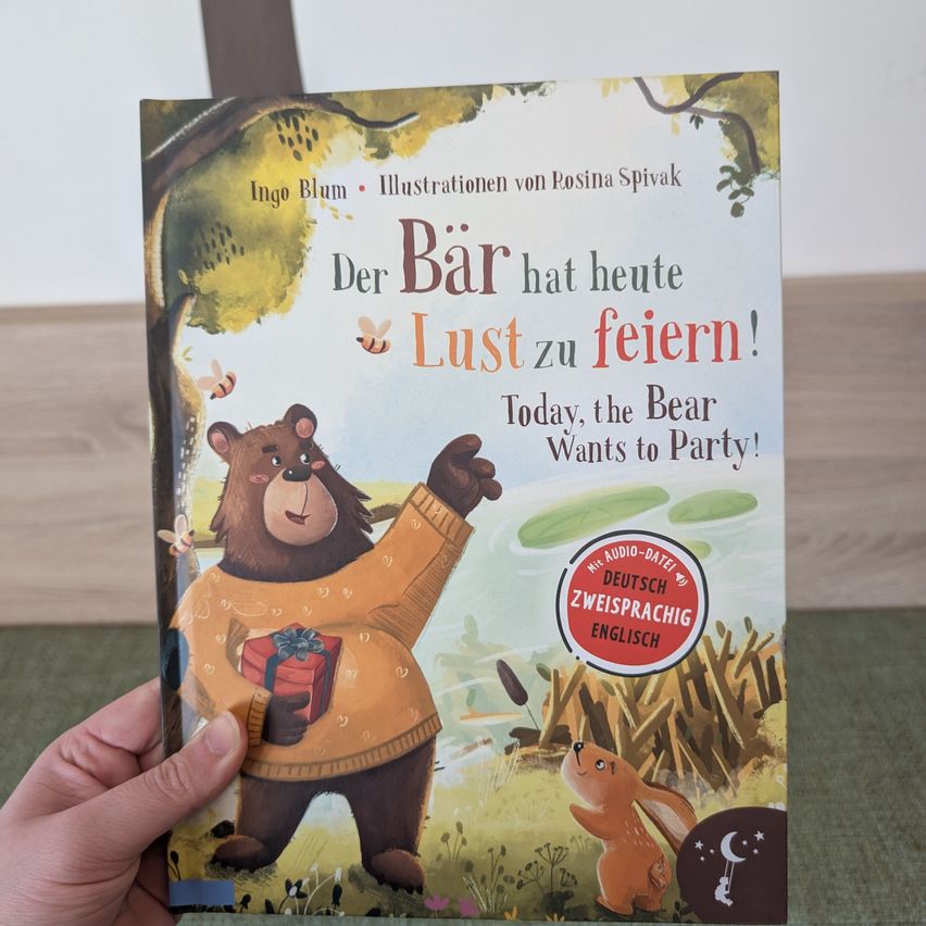 Ein Buchcover zeigt einen Bären, der ein Geschenk hält, mit dem Text 'Der Bär hat heute Lust zu feiern!' und einem Aufkleber für ein Audio-Datum in Deutsch und Englisch.