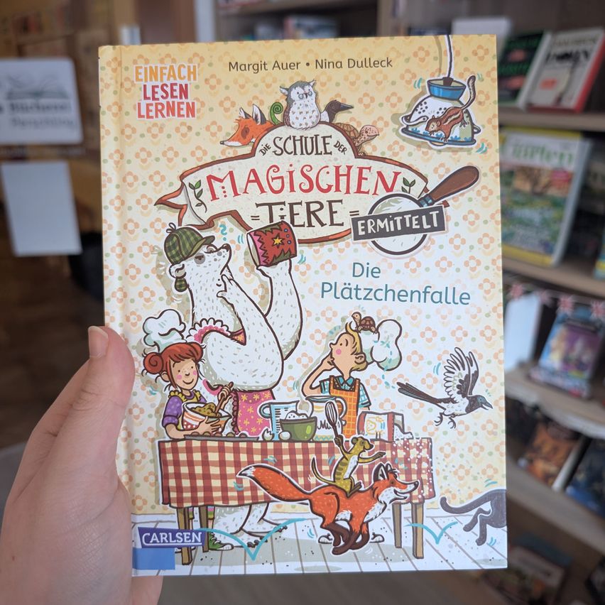 Eine Hand hält ein Buch mit dem Titel 'Die Plätzchenfälle'. Das Cover zeigt Illustrationen von Kindern, Tieren und einer Küchenszene. Es ist Teil der 'Schule der magischen Tiere'-Reihe.